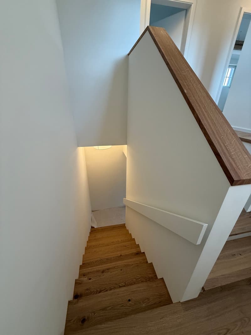 Wooden staircase with white walls and a wooden handrail leading to a room above.