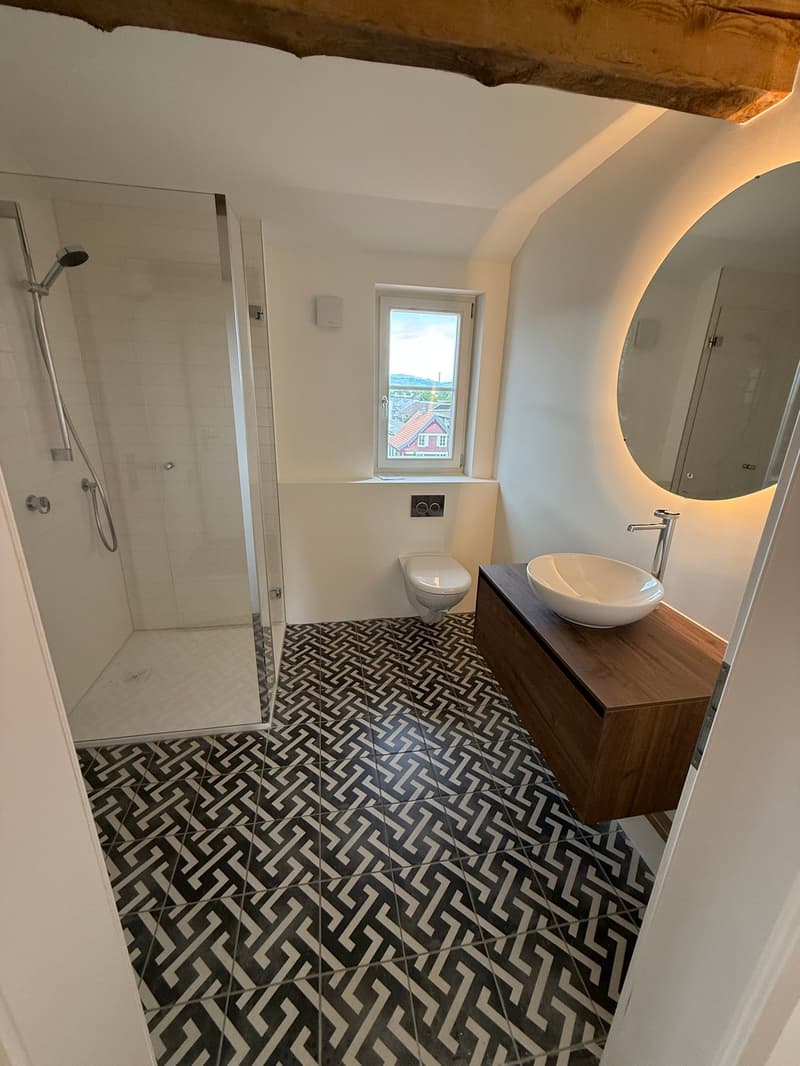 The image shows a modern bathroom with a patterned tile floor, a vanity with a vessel sink, a large mirror, and a glass shower enclosure. The bathroom has a clean, minimalist design with white walls and wooden accents.
