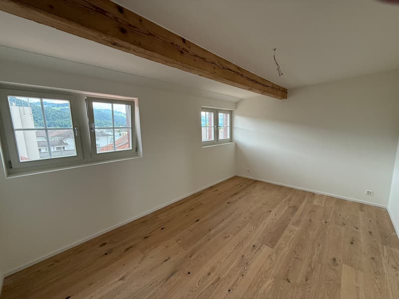 Spacious bedroom with hardwood floors, white walls, and exposed wooden beams on the ceiling. Large windows provide natural light and a view of the surrounding area.