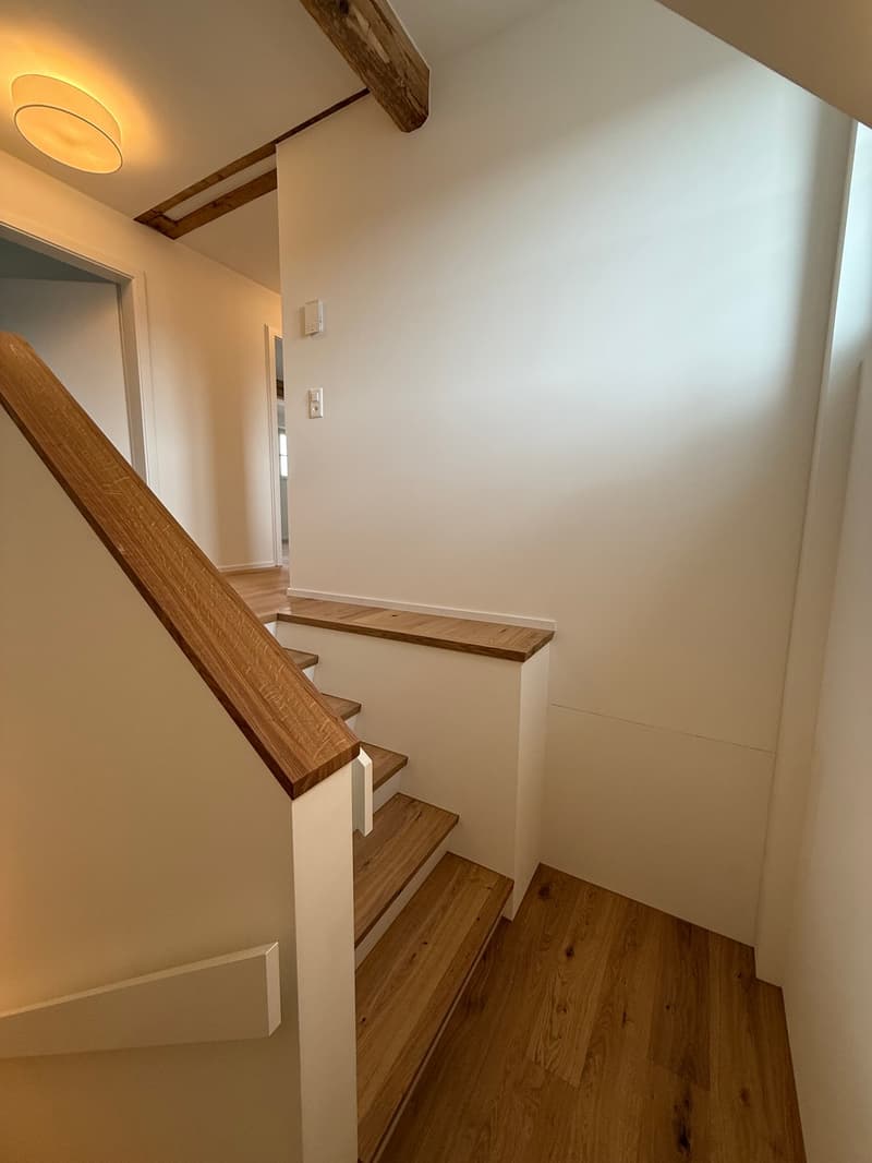 Wooden staircase with white walls, wooden beams on the ceiling, and a wooden handrail.