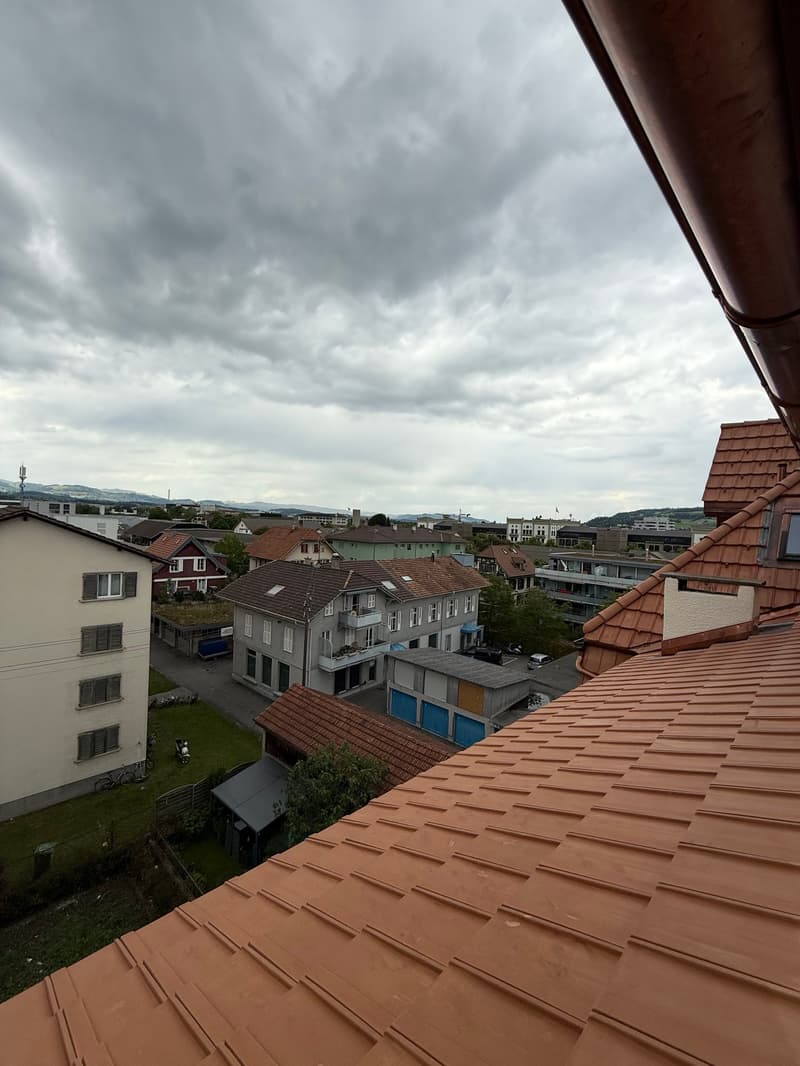 The image shows a view of a residential neighborhood with a mix of multi-story buildings and houses with red tile roofs. The sky is overcast with dark clouds, creating a gloomy atmosphere. The buildings appear to be located on a hillside, with some struct