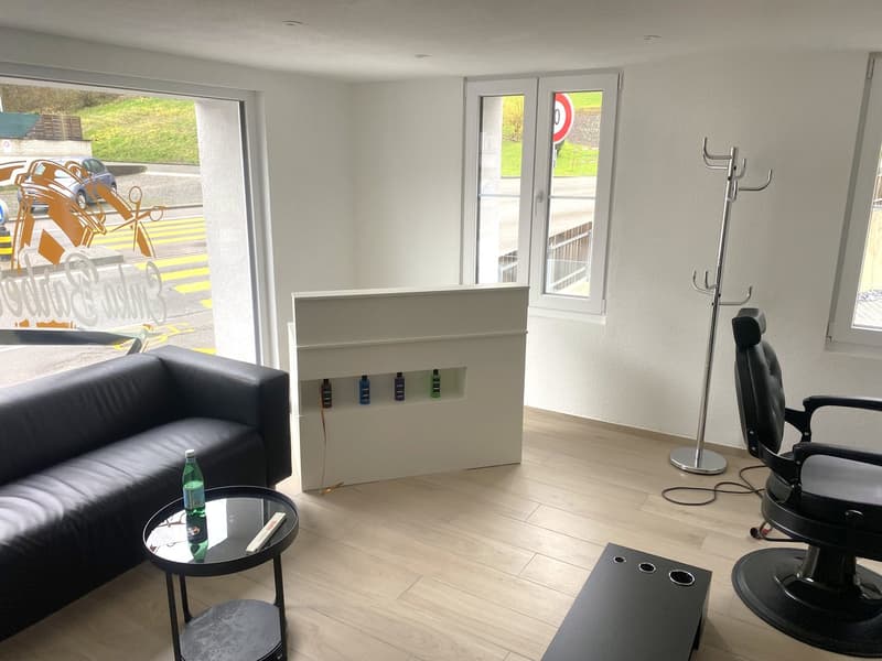 Spacious living room with large windows, hardwood flooring, black leather sofa, coffee table, and a white storage unit with shelves for displaying items.