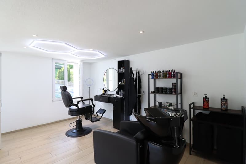 The image shows a hair salon with a modern and minimalist design. The room has white walls and a tiled floor. There is a large window that provides natural light and a view of the outdoors. The salon features black salon chairs, a styling station with a m