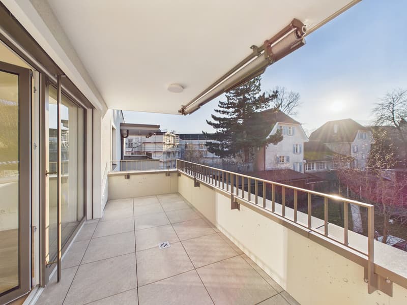 A wide balcony with sliding glass doors, metal railings, tiled flooring, and a view of buildings and trees