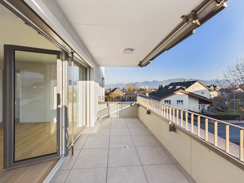 A balcony with tiled flooring and a railing. The balcony has large glass doors. The balcony has a view of the surrounding area, including houses and mountains.