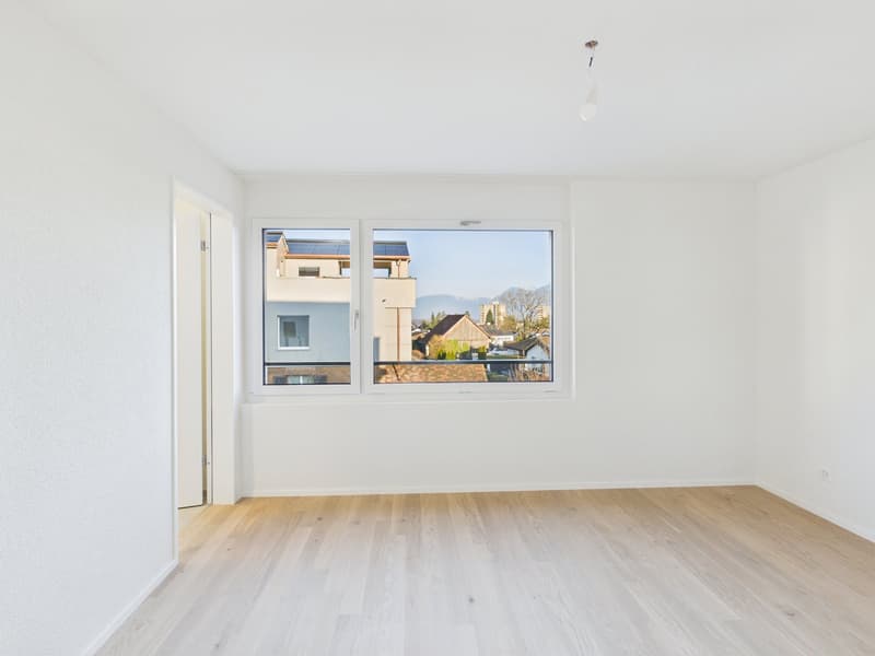 Empty room with white walls, wooden floor, large window with outdoor view.