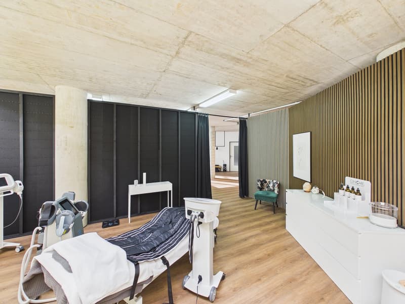 Examination room with wooden floor, concrete ceiling, treatment bed, medical equipment, cabinets, and a stool