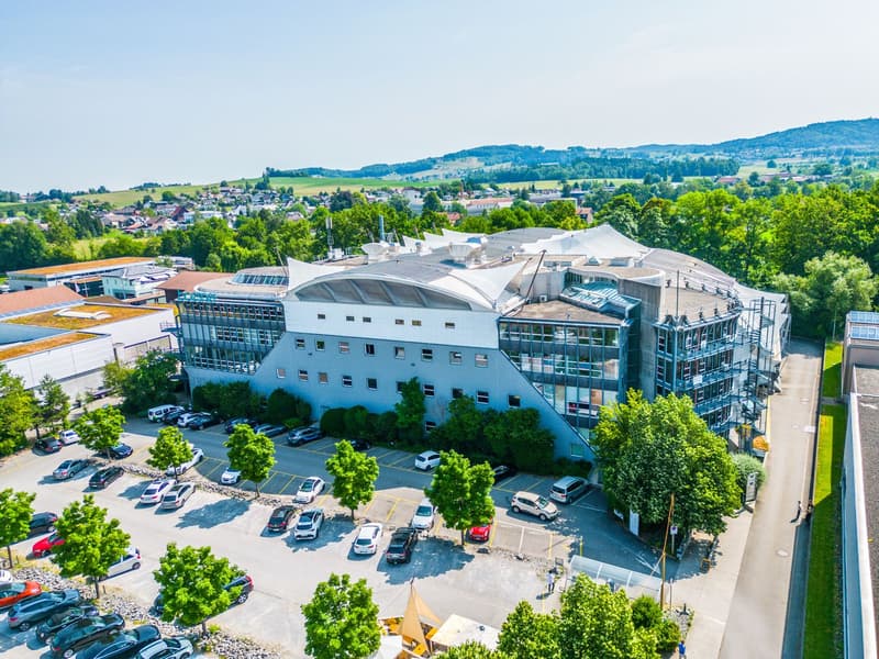 Modern architectural building, glass facade, several levels, surrounded by parking lots, greenery