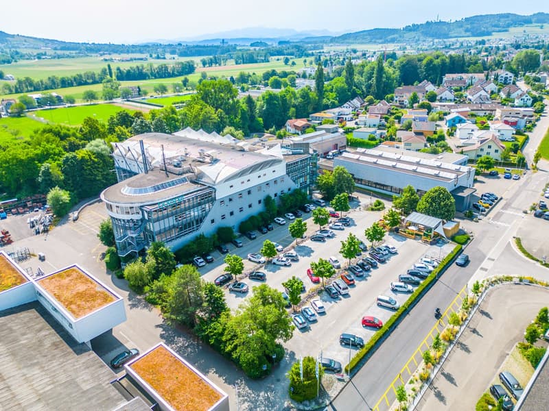 A large building with several wings, surrounded by trees and a large parking lot, houses and trees in the background