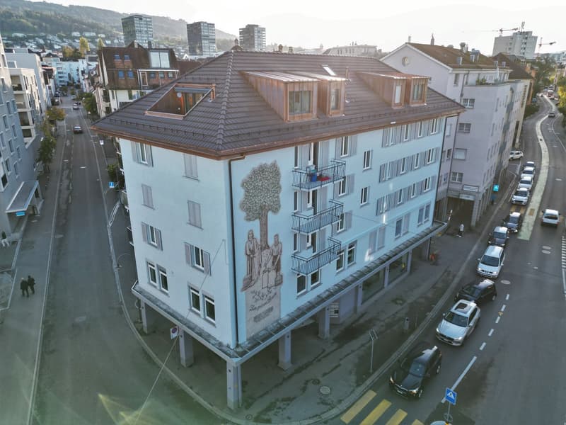 A multi-story building with a brown roof, white walls, and a mural on the side. It has multiple windows and balconies. The building is situated on a raised ground floor with pillars supporting it.