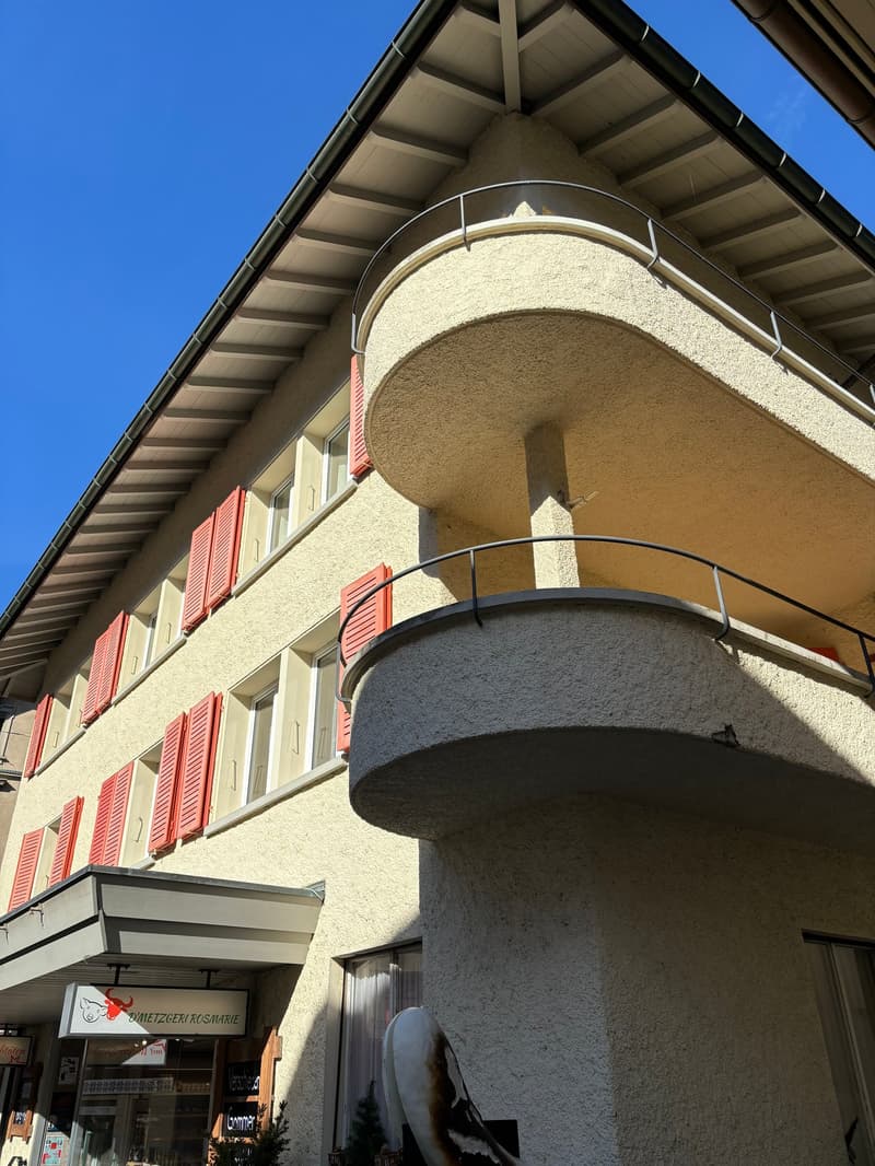 2-story building with a curved facade, beige exterior walls, red shutters on the windows, and a balcony on the upper level.