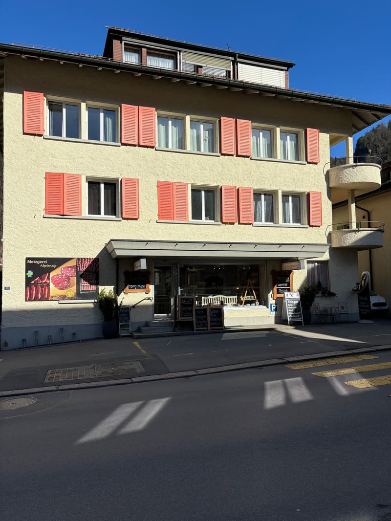 2-story building with yellow exterior walls, red shutters, and a balcony on the upper floor. The ground floor has a storefront with a meat market sign.