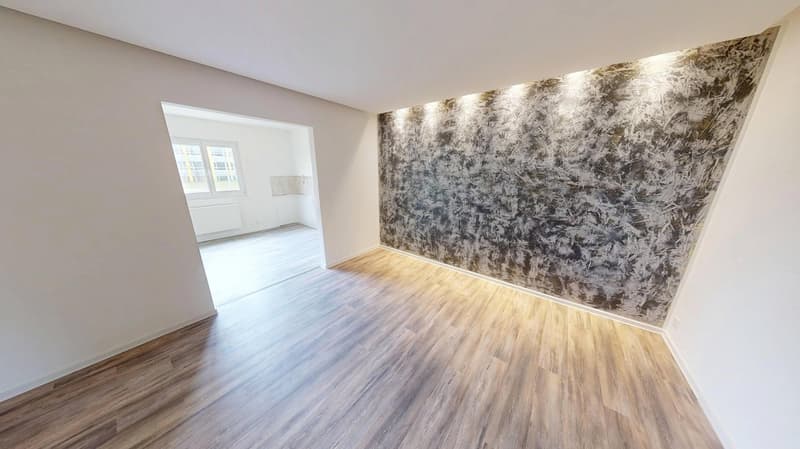 Empty room with hardwood floors, an entrance to a small hallway, and a window with a white frame.