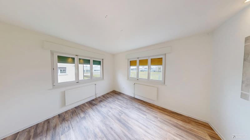 Empty room with wooden floors, two large windows, and white walls.