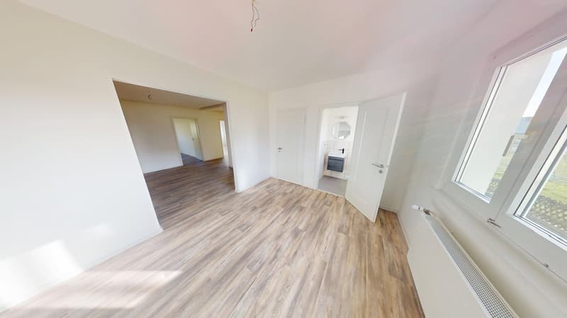 Empty room with white walls, wooden floors, open door, small window, mirror on the hallway, white radiator, bathroom glimpse