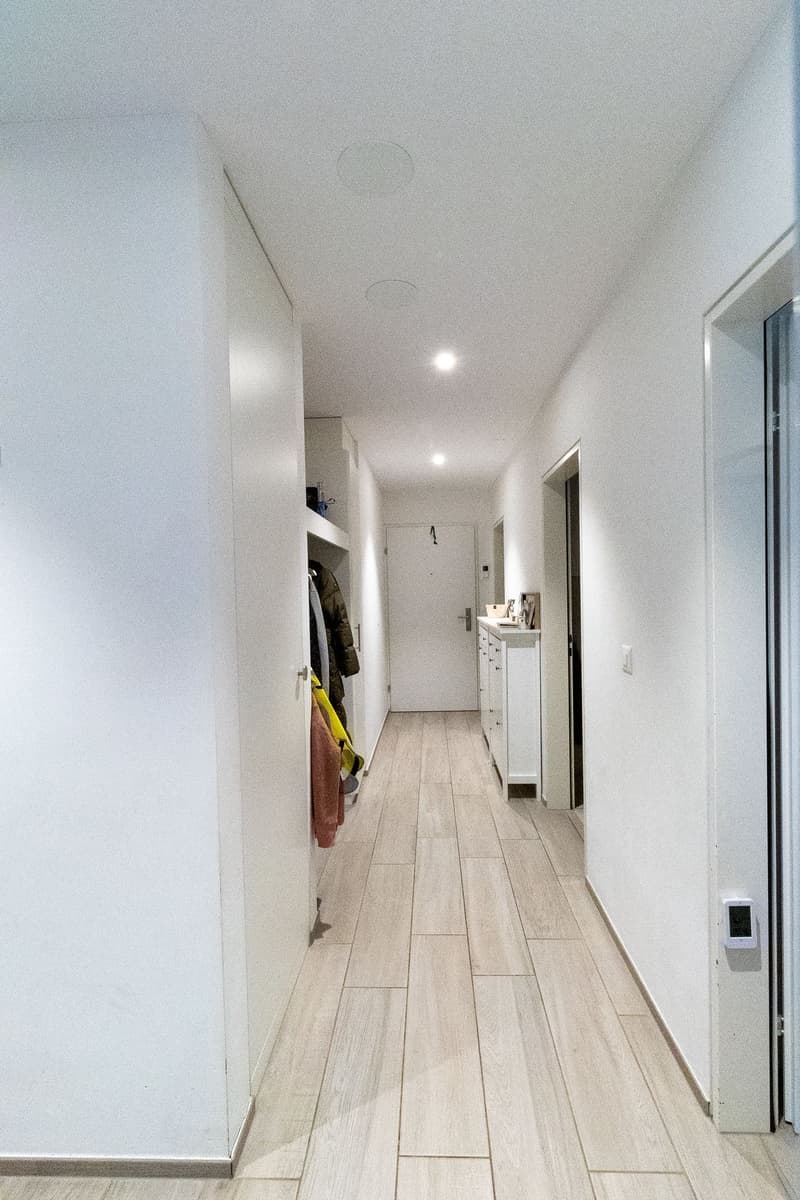 Empty hallway with white walls, wooden floor, and ceiling lights. There are two doors, one of which leads to a room with a white cabinet and a coat rack.