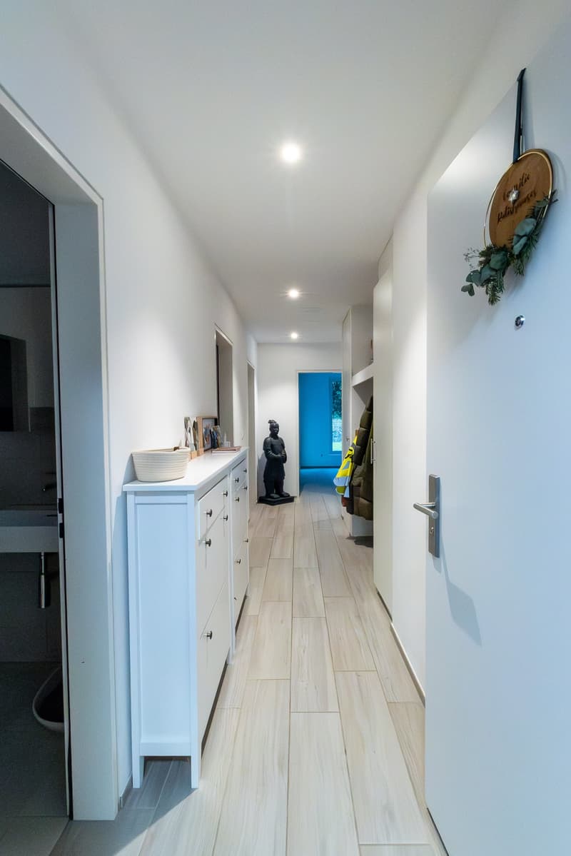 A hallway with white walls and wooden floor, a white cabinet with drawers, a statue, and a wreath on the wall.