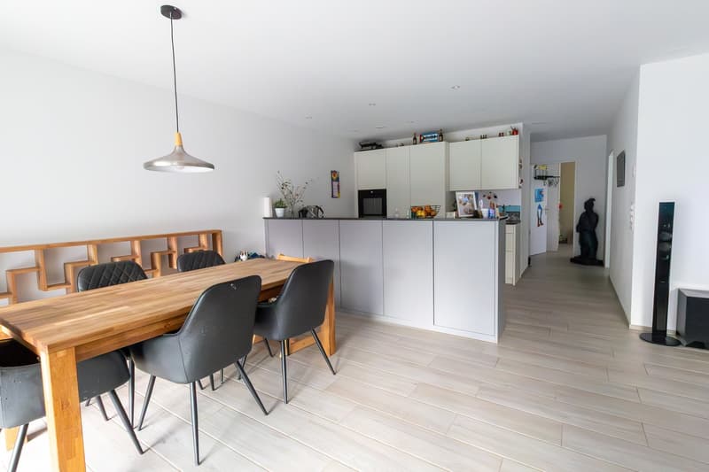 Dining room with wooden table, gray chairs, white walls, wooden floor, kitchen with white cabinets, microwave, refrigerator, and a statue