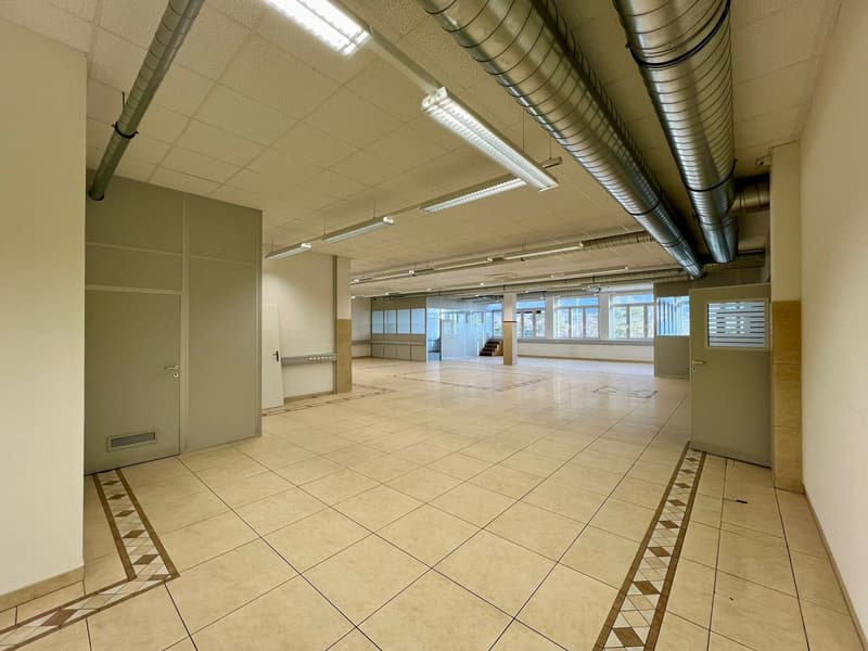 The image shows a large, open commercial space with a tiled floor and exposed ceiling ductwork. The space appears to be an office or retail area, with a reception desk or counter visible in the background. The room has ample natural light coming in throug