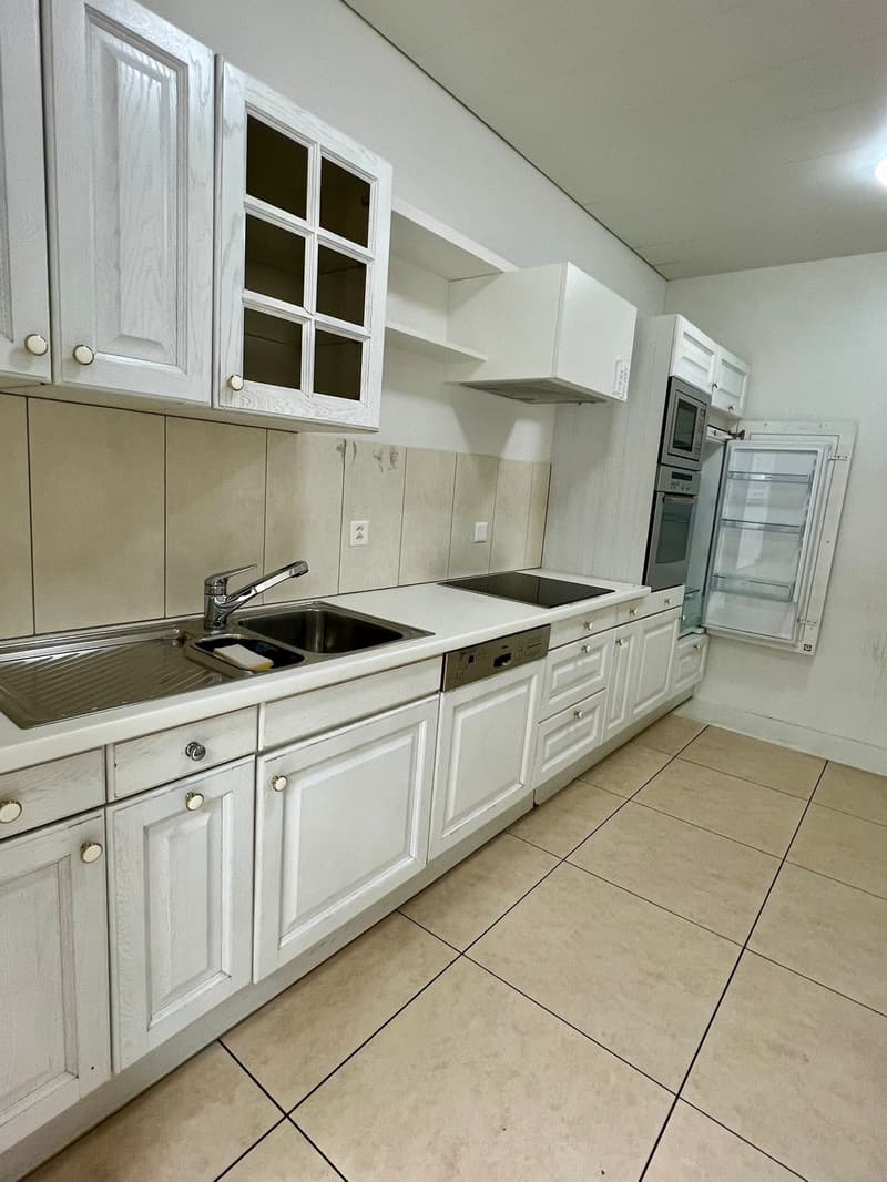 The image shows a spacious kitchen with white cabinets, a sink, and a stove. The kitchen has tile flooring and appears to be well-equipped, including a dishwasher.