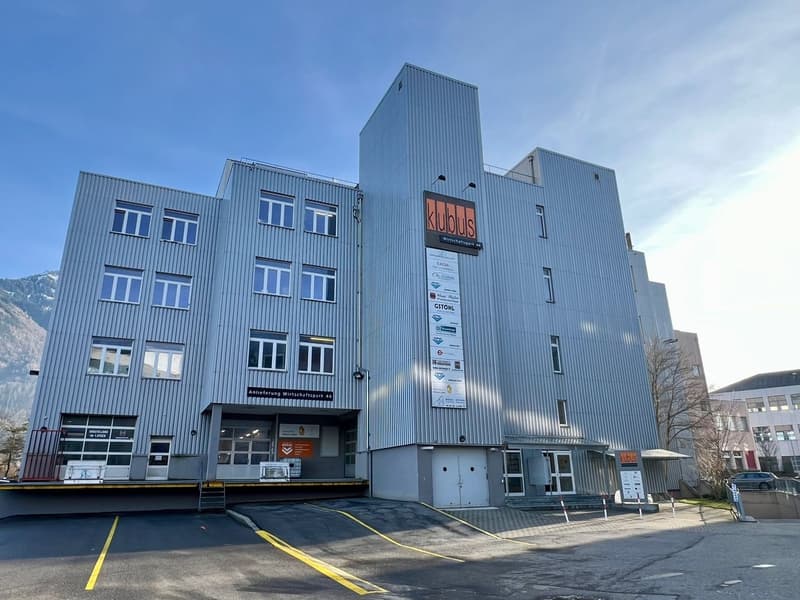 A multi-story commercial building with a gray metal facade and large windows. The building has the name 'Klubus' displayed prominently on the front. There are parking spaces in front of the building.