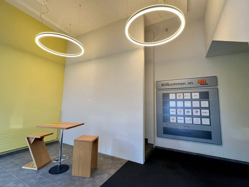 The image shows an office lobby or reception area. The space features modern, minimalist design elements such as circular pendant lights, wooden benches, and a digital check-in kiosk with the text 'Willkommen im kudus' (Welcome to kudus). The walls are pa