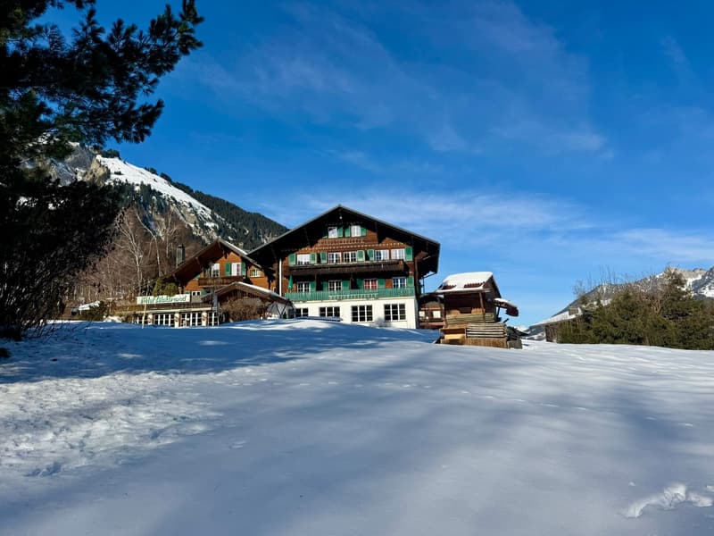 Chalet-style house, snow-covered exterior, surrounded by snow and pine trees, mountainous background, multiple buildings in the complex