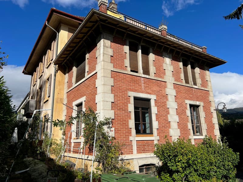 2-story historic brick building with stone accents, large windows, and a sloped roof. The building appears to be surrounded by greenery and vegetation.