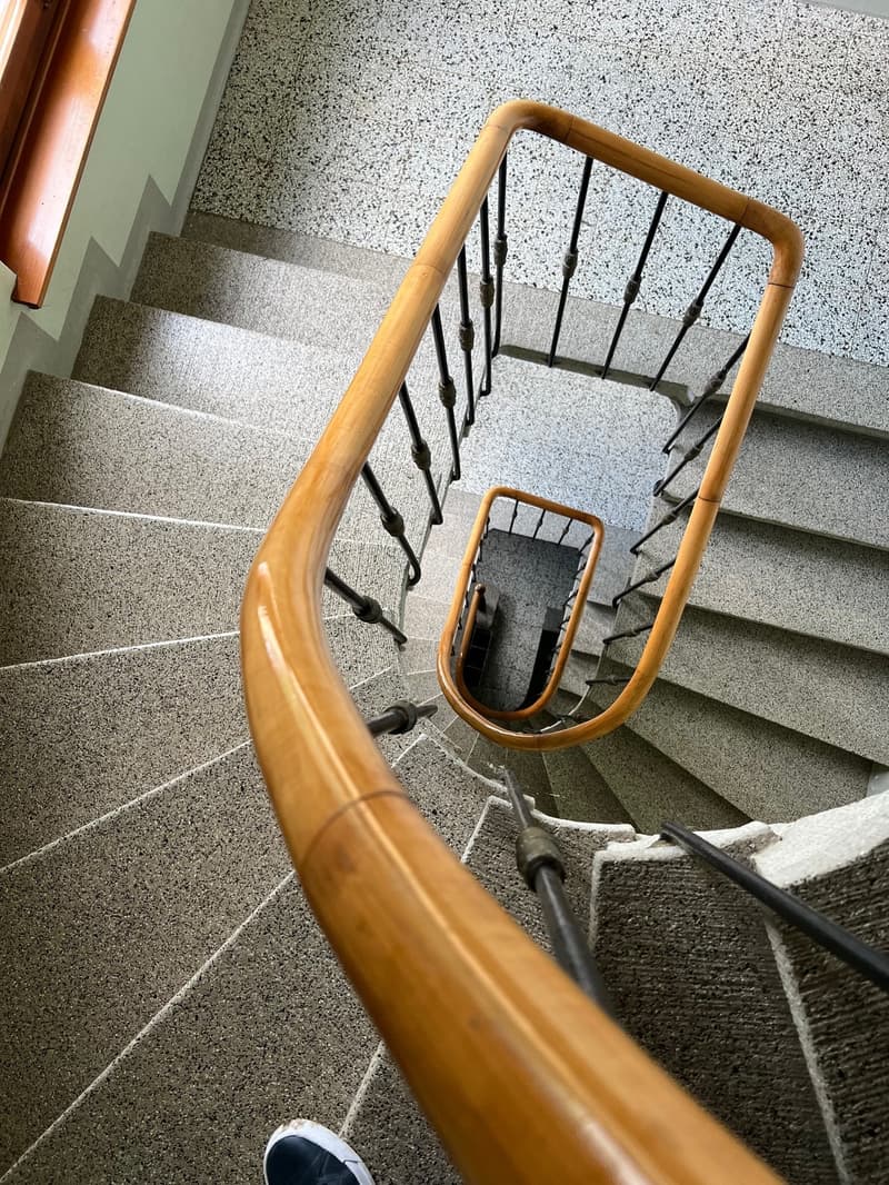 Spiral staircase with wooden handrail and metal railings, leading up to a terrazzo floor landing
