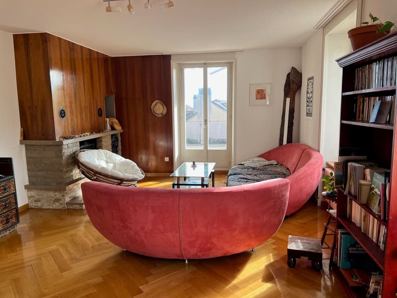 Spacious living room with hardwood floors, wooden wall paneling, large windows, red velvet sofa, glass coffee table, and a fireplace. The room also features a bookshelf, potted plant, and various decor items.