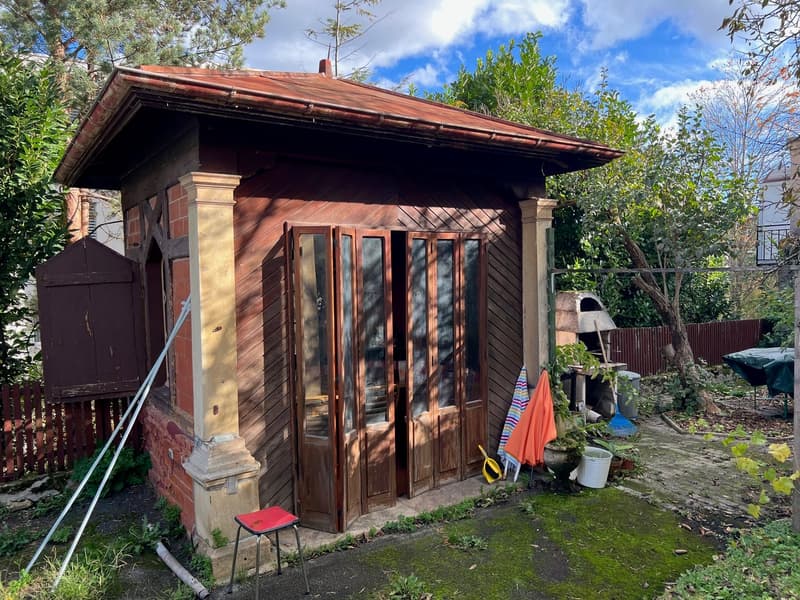 A wooden shed with a red roof, wooden walls, and large windows. The shed is located in a garden setting with trees and other plants surrounding it.