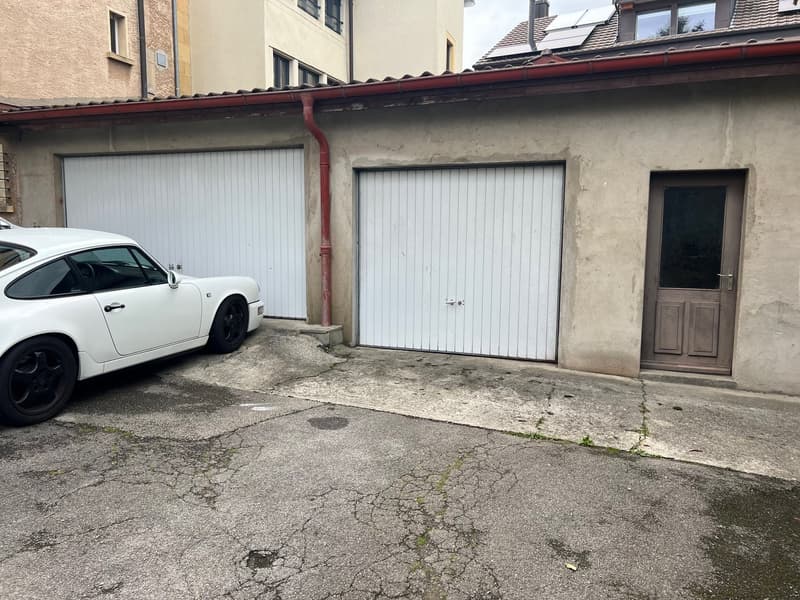 The image shows a two-story building with a white car parked in front. The building has a garage with two garage doors and a separate entrance door. The exterior walls appear to be made of stucco or a similar material, and the roof is tiled. The ground in
