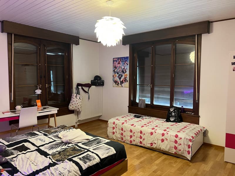 The image shows a bedroom with a large bed, a desk, and a white feather chandelier. The room has wooden furniture and windows, and the walls are decorated with artwork and posters.