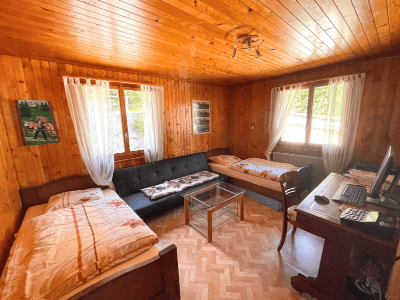 room with wooden walls, 3 beds, couch, desk, computer, glass table, radiator, windows with curtains