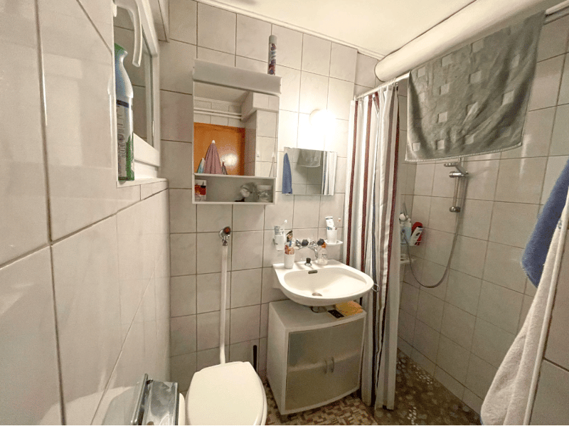 white tiled walls, white sink, white toilet, shower with curtain, mirror, towel rack, small cabinet under sink