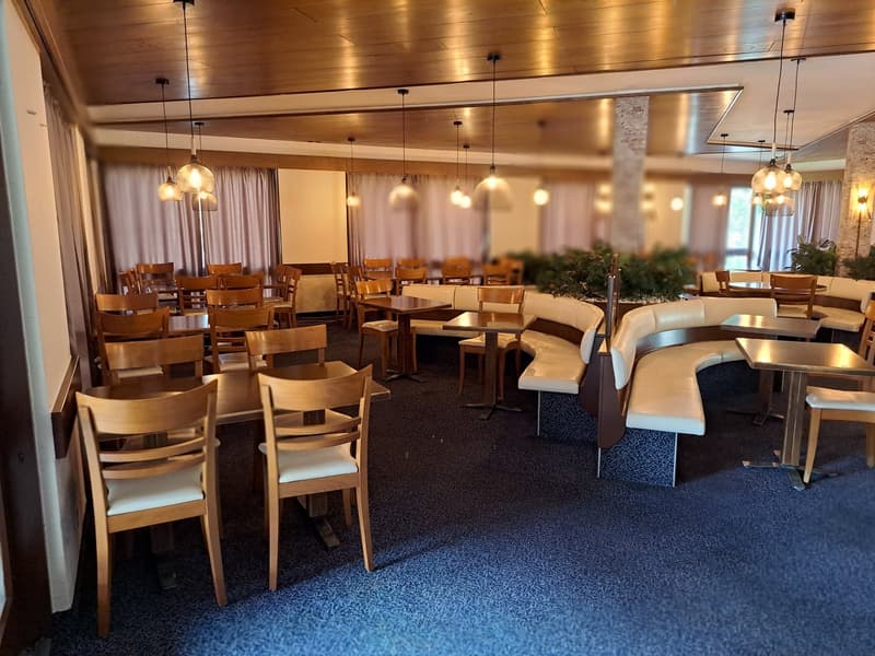 The image shows a spacious dining area with wooden tables and chairs, as well as comfortable upholstered seating. The room has a warm, cozy atmosphere with wooden paneled walls and ceilings, and pendant lighting fixtures. The floor is covered in a blue pa