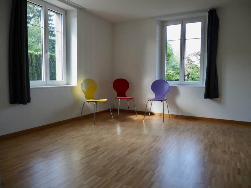 room with windows, wooden floor, three colorful chairs, white walls