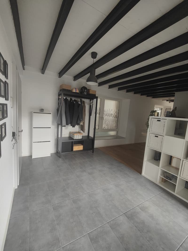 Empty room with gray tiles, white walls, black beams, white cabinet, clothes rack, baskets, lamp, framed pictures, door