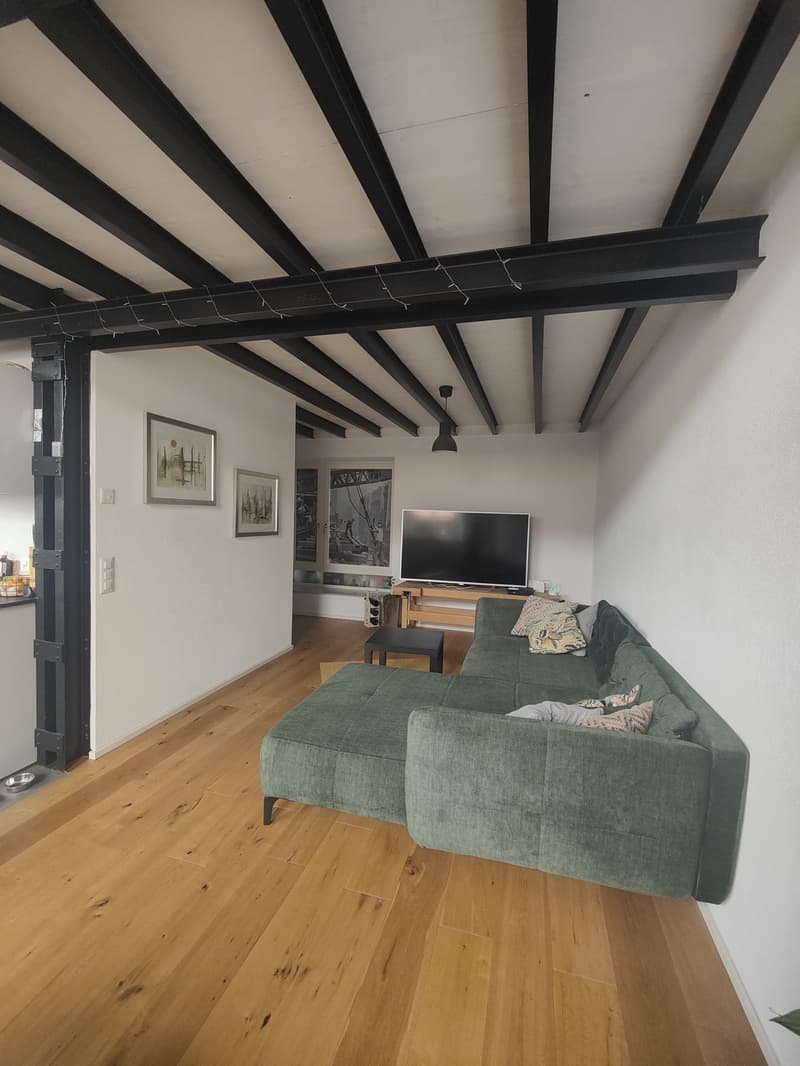 Living room with wooden floor, black beams, green sofa, TV, coffee table, picture frames, and a lamp