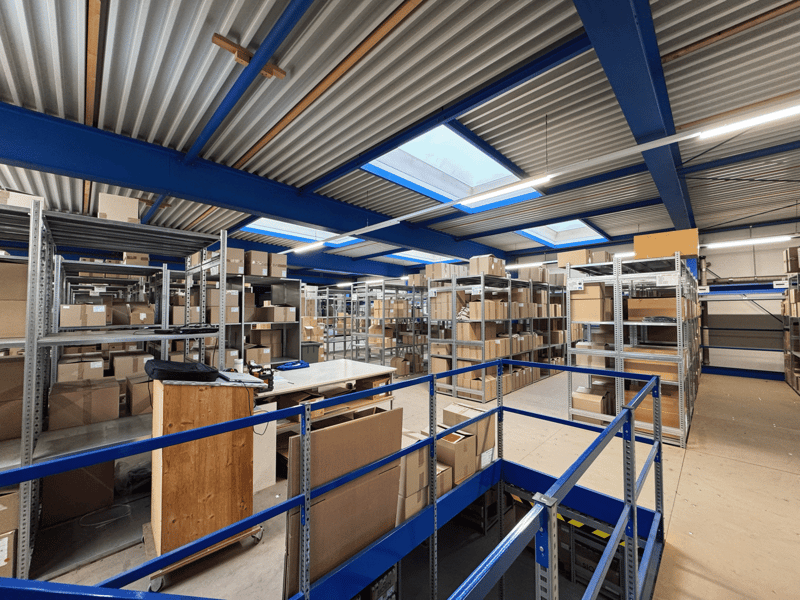 Large storage area with multiple racks and shelves, filled with boxes. Metal blue railings, blue and silver ceiling beams, daylight coming in through several skylights