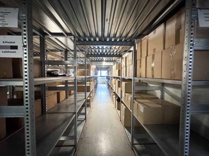 Long hallway with steel shelves on both sides filled with brown boxes, metal ceiling, and white floor.