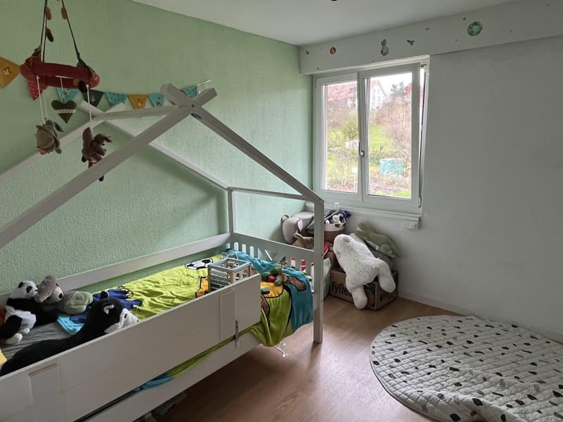 Children's bedroom with a bed, toys, storage box, decorative toys, green walls, white walls, floor, round rug, window with an outdoor view