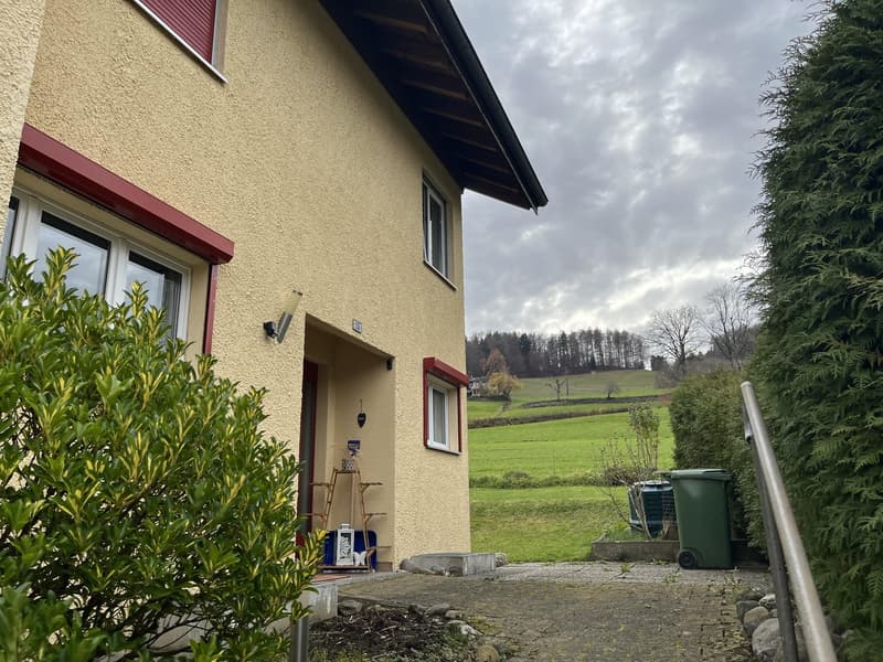 yellow stucco exterior, red trims, three windows, green grass in the background, trash bin