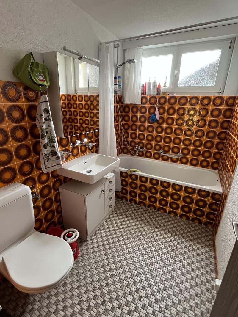 Bathroom with white toilet, a bathtub, a sink with a cabinet, shower with a handheld head. The room has checkered tiles and a patterned wall.
