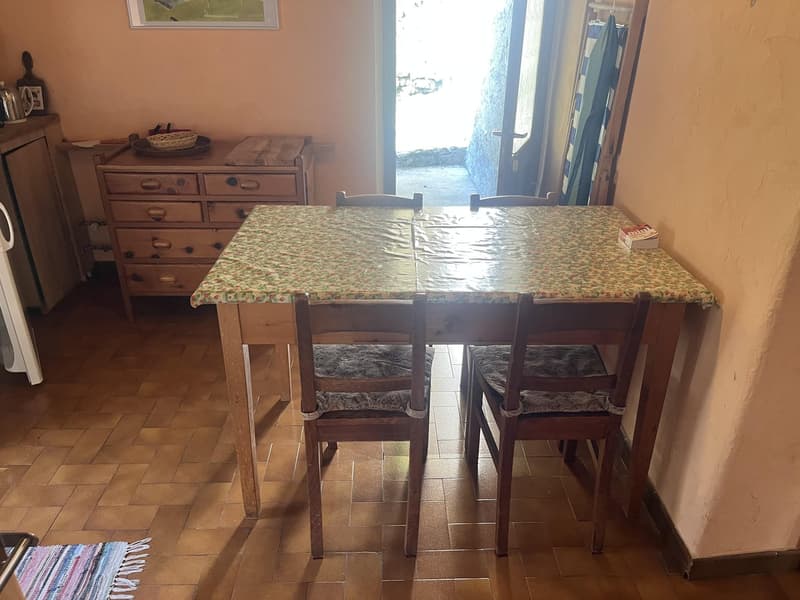 wooden table with 4 chairs, floral placemats, wooden drawer, books, white bucket on the cabinet, an open door with light