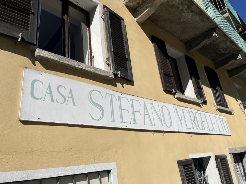 A yellow building with a sign that says 'CASA STEFANO VERGELETTO'. It has several windows with wooden shutters, and a balcony with metal railings on the top floor.