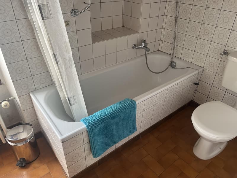 Bathtub, white tile walls, floored in wood, blue towel on the tub, toilet, waste basket, no windows.