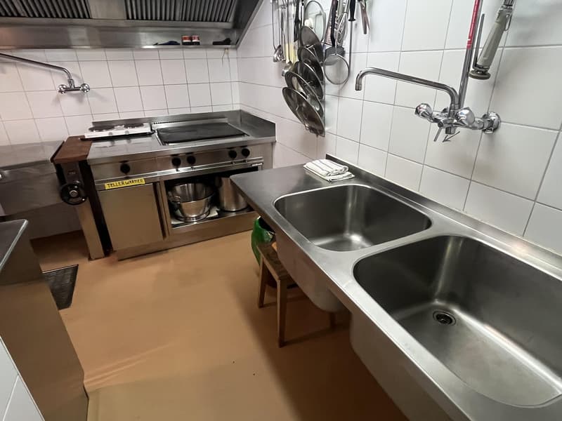 Stainless steel countertops, gas stove, double sink, hanging pots and pans, tiled wall, wooden stool