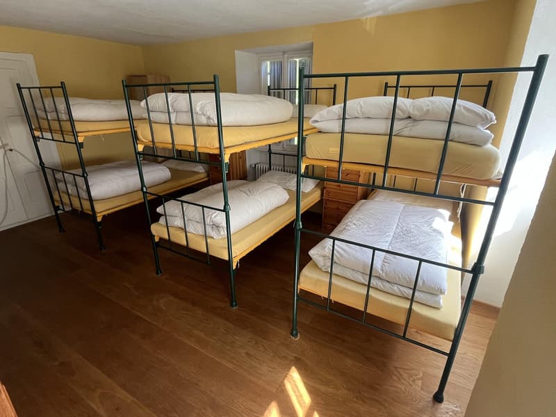 Four bunk beds, each bed has a yellow mattress, white sheets, pillows, wooden floor, white walls, two windows, radiator
