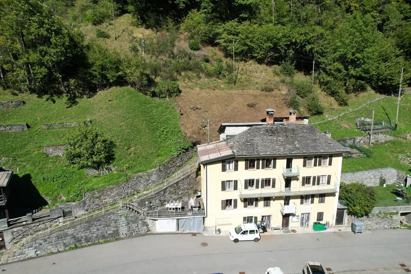 yellow building, multiple windows, balcony, stone wall, parking area, trees, grass